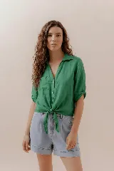 Camisa verde de lino, con cuello camisero, mangas cortas remangadas y cierre frontal de botones. Presenta un lazo en la parte inferior para anudar en la cintura.