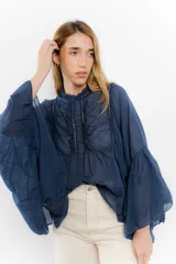 Camisa de algodón con diseño romántico, cuello alto con volados y detalles de puntillas verticales en el frente. Presenta mangas largas acampanadas con gran volumen y corte holgado.