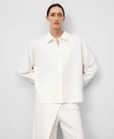 Camisa blanca de manga larga con cuello solapa y botonadura frontal. Presenta un bajo redondeado y un pliegue en el canesú de la espalda.