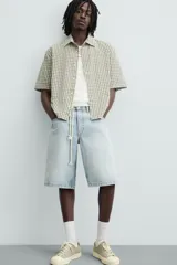 Bermuda de denim de algodón, color azul medio lavado, de corte ancho (baggy fit) y largo hasta la rodilla. Presenta cinco bolsillos y cierre frontal con cremallera y botón.