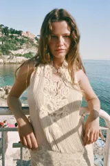Vestido midi de color beige con escote en pico y tirantes anchos. Presenta un diseño con tejido plisado en la parte superior y aplicaciones de encaje a tono en el escote y la cintura. Cuenta con una tira ajustable con lazada en la espalda y forro interior.