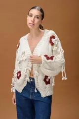 Saco de punto grueso color crudo, con diseño de calce relajado y mangas amplias. Presenta apliques tejidos con motivos florales en color terracota y detalles de tiras anudadas decorativas en toda la prenda.