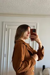 Campera tipo bomber de corte corto, confeccionada en gamuza color camel. Presenta cuello solapa, cierre frontal con botones, forro interior y detalles decorativos de pasamanería trenzada a lo largo de las mangas.