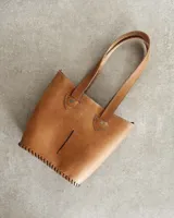 Bolso tipo tote de cuero color marrón claro, con doble asa de hombro, detalle de costuras a la vista y abertura frontal.