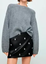 Minifalda de corte recto efecto piel, color negro, con diseño corto y adorno de tachuelas metálicas repartidas por la superficie. Cuenta con cierre de cremallera lateral y forro interior.