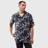 Camisa de manga corta con cuello cubano, confeccionada en viscosa. Presenta un estampado botánico de hojas en tonos grises y azules sobre fondo negro, con detalles de texto manuscrito integrado en el diseño. Incluye un bolsillo frontal en el pecho y cierre de botones.