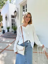 Cartera tipo sobre color crudo, confeccionada en cuero, con correa larga de cuero marrón y costuras decorativas en contraste.