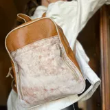 Mochila de cuero color camel con textura tipo piel de reptil en la parte superior y diseño irregular en blanco y marrón en la parte inferior. Tiene cuatro compartimentos independientes con cierre, dos bolsillos internos y correas de cuero regulables.