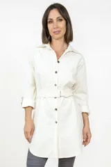 Sobre camisa de corte largo y estilo minimalista, color blanco, con cuello camisero, cierre frontal de botones y cinturón ajustable a la cintura. Presenta mangas largas con puños y bolsillos laterales.