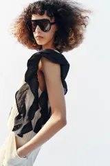 Blusa de cuello redondo y manga sisa, de color beige con rayas verticales. Presenta volantes negros en las sisas y una tira de encaje central con jaretas. Cuenta con cierre en la espalda con abertura y botón.