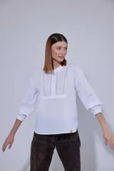 Camisa blanca de algodón con pechera decorada con alforzas y puntillas. Presenta cierre frontal con botones, mangas tres cuartos con frunces y detalles de puntilla, y un diseño de corte holgado con ruedo redondeado.