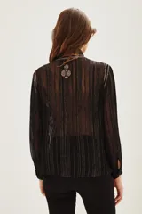 Camisa de manga larga en color negro con tejido semitransparente y detalles de hilos metalizados. Presenta un diseño con bordados florales en tonos tierra, cierre frontal con botones y un lazo decorativo en el cuello.