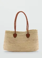 Bolso tipo cesta de playa confeccionado en hoja de palma trenzada, con doble asa de hombro larga en cuero trenzado color marrón y detalles de refuerzo en cuero en la base de las asas.