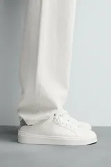 Zapatilla deportiva de cuero sintético color blanco, con suela de plataforma gruesa y detalle de gamuza gris en el talón.