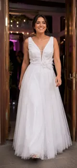 Vestido largo de fiesta color blanco, confeccionado en tul bordado con hilos y brillos. Presenta escote profundo en V con transparencia central, corte en la cintura y apliques decorativos de flores en tela, plumas y perlas.
