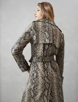 Trench de ecocuero con estampado animal print de serpiente en tonos beige, marrón y negro. Presenta cuello con solapa ancha, doble abotonadura frontal con botones negros, cinturón ajustable con hebilla metálica, mangas largas con presillas y botones en los puños, canesú suelto en la espalda, bolsillos laterales y forro interno.