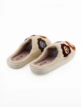 Pantuflas de estar por casa tipo mule, confeccionadas en tejido de peluche color crema. Presentan un diseño con apliques bordados de caritas sonrientes en tonos marrones, bordó y lila. Cuentan con una suela plana de goma y forro interior suave.