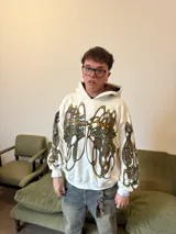 Hoodie oversize de color blanco con estampado artístico en tonos marrones y dorados en el pecho y las mangas. Presenta un forro interior de capucha en tejido marrón texturizado y costuras reforzadas.
