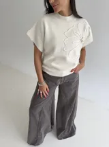 Chaleco tejido de punto color crudo, con cuello redondo y diseño de flor en relieve en el frente. Presenta un corte holgado y mangas cortas con terminación acanalada.