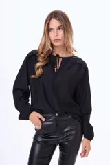 Blusa negra de corte holgado con mangas largas abullonadas, puños elásticos y detalle de volados en los hombros. Presenta un escote redondo con abertura frontal y lazos para anudar.