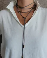 Collar tipo corbatín confeccionado en tiento de cuero gris, con múltiples vueltas alrededor del cuello y detalle de pequeñas esferas metálicas en tono bronce distribuidas a lo largo de la tira.