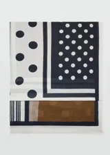 Pañuelo fluido de tejido ligero, con diseño estampado que combina secciones en blanco con lunares negros, un panel en negro con lunares blancos, y una franja inferior en marrón y gris con detalles de rayas. Presenta terminaciones deshilachadas en los bordes.