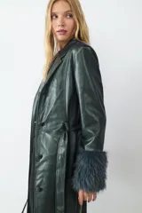 Trench largo de efecto cuero con acabado brillante, cuello solapa, cierre frontal con botones y cinturón ajustable a la cintura. Presenta puños con detalle de pelo sintético y diseño estructurado.