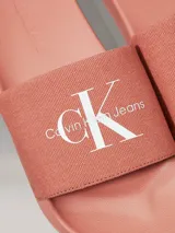 Ojotas tipo slide de color rosado, con base de EVA y correa ancha de lona de algodón reciclado. La correa presenta el logo de Calvin Klein Jeans bordado en blanco.