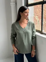 Blusa de manga larga en color verde oliva, con cuello en V y costuras decorativas visibles. Presenta un corte recto y holgado con un ligero fruncido en los hombros.