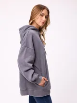 Canguro oversize de color gris con efecto lavado, capucha con cordones ajustables, bolsillo frontal tipo canguro y parche pequeño con logo de la marca en el pecho.