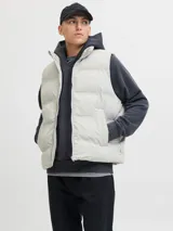 Chaleco puffer acolchado de color gris claro, con cuello alto y cierre frontal con cremallera. Cuenta con bolsillos laterales y diseño versátil sin mangas.