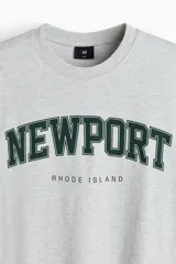 Remera de algodón gris jaspeado con cuello redondo y mangas cortas. Presenta un estampado frontal con la inscripción 'NEWPORT' en tipografía universitaria verde oscuro y 'RHODE ISLAND' debajo.