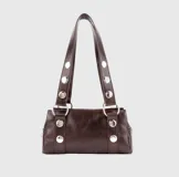 Bolso de hombro pequeño color marrón oscuro con acabado brillante, estilo baguette. Las asas son ajustables y están decoradas con ojales metálicos plateados.