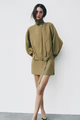 Vestido corto verde militar de cuello solapa y manga larga, con bolsillos de plastrón en el delantero y cierre frontal con cremallera oculta por sopata. Cuenta con un cinturón incorporado del mismo tejido con hebilla metálica y aberturas laterales en el bajo.