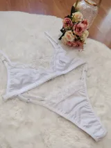 Conjunto de lencería compuesto por un bralette triangular y una tanga, ambos confeccionados en algodón blanco con detalles de encaje en los bordes. El bralette cuenta con breteles ajustables y la tanga presenta laterales regulables.