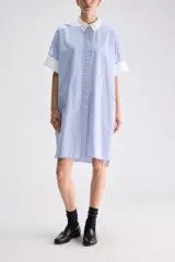 Vestido camisero midi de calce relajado y silueta fluida, confeccionado en tejido a rayas celestes y blancas. Presenta cuello clásico y puños en contraste de color blanco, mangas cortas con vuelta y cartera frontal oculta con botones.