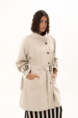 Trench corto color beige con cuello alto, cierre frontal de botones y cinturón ajustable en la cintura. Presenta mangas largas con detalles de tiras ajustables en los puños y bolsillos laterales.