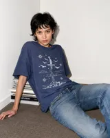 Remera oversize de algodón con estampa frontal en color blanco que representa una espada clavada en una roca, rodeada de estrellas, nubes y aves.