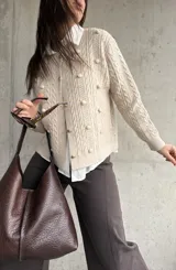 Cardigan de punto color beige con diseño de trenzas y pompones decorativos. Presenta cuello redondo, cierre frontal con botones y mangas largas.
