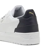 Championes urbanos Puma modelo Shuffle Downtown, de diseño clásico con base blanca y detalle en azul marino en el talón. Presenta el logo de la marca en dorado en el lateral y la lengüeta, con suela de goma blanca y cordones al tono.