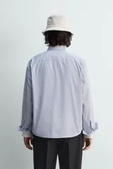 Camisa de algodón de corte relajado, con cuello solapa abotonado (button-down) y manga larga terminada en puño con botón. Presenta un patrón de rayas verticales finas en tonos azul claro y blanco.