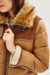 Campera estilo aviador en tonos tostados, con diseño acolchado y paneles de gamuza en la parte superior. Presenta cuello alto con doble hebilla, cierre frontal completo y detalles de corderito sintético en el cuello y el ruedo.