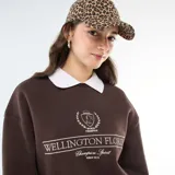 Buzo de felpa color marrón con cuello polo blanco en contraste. Presenta un estampado gráfico frontal con estilo universitario que incluye la inscripción 'Wellington Florida' y un emblema central.