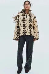 Buzo oversized de sherpa suave en color beige con estampado geométrico en tonos marrones y detalles en tejido revestido. Presenta cuello elevado, medio cierre frontal y bolsillos laterales.