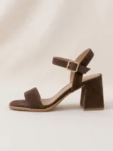 Sandalias de gamuza color marrón chocolate, de taco cuadrado medio y punta cuadrada. Tienen una tira delantera ancha y una tira ajustable al tobillo con hebilla metálica dorada.