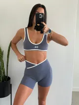 Conjunto deportivo de dos piezas en color gris azulado con detalles de ribete blanco. Incluye un top de escote redondo con logo estampado y un short de tiro alto con diseño cruzado en la cintura.