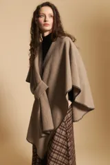Poncho de tejido suave color beige con diseño envolvente y terminaciones con costuras decorativas en contraste color negro. Presenta un detalle de lazo frontal integrado.