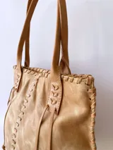 Cartera tipo tote de cuero color camel, con diseño artesanal que presenta costuras decorativas visibles en los laterales y el frente. Cuenta con doble asa de hombro y cierre superior.