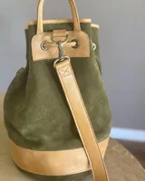 Mochila tipo bucket de gamuza marrón, con detalles de cuero liso en el tono, bolsillo frontal con cierre y cordón ajustable con tiras.