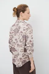 Camisa de manga larga con cuello clásico y cierre frontal de botones. Presenta un estampado floral en tonos marrones sobre una base color beige, confeccionada en un tejido con textura similar al gamuzado.